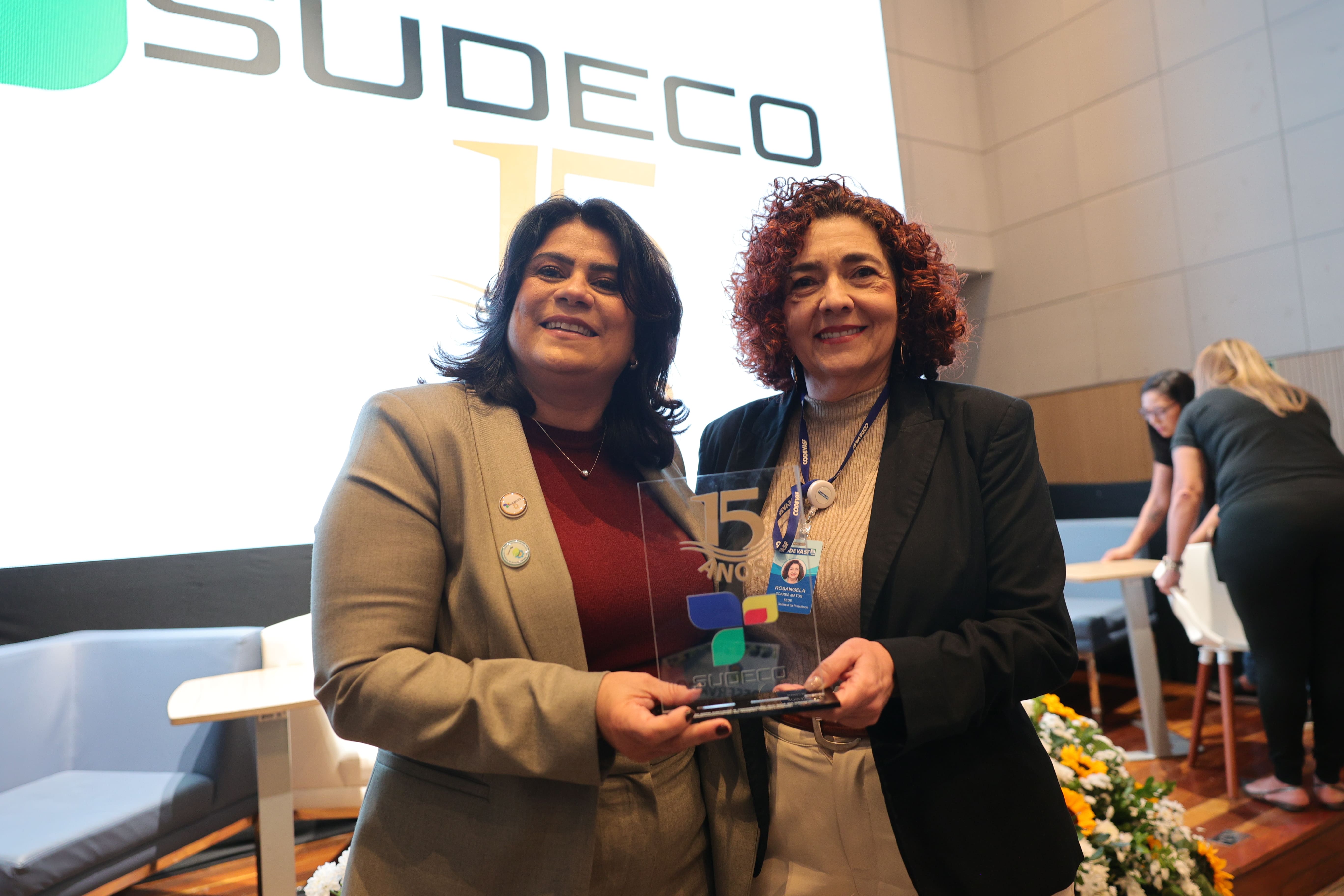 Duas mulheres seguram placa comemorativa em evento com painel da Sudeco ao fundo