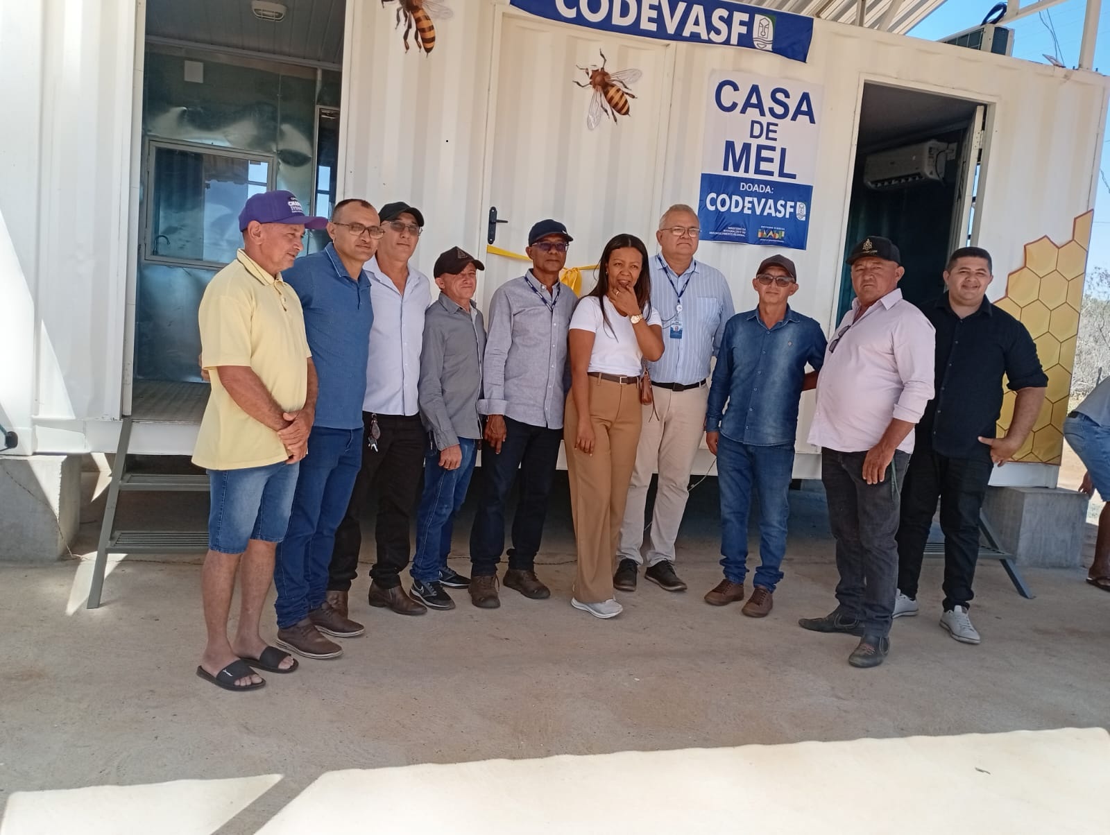 Grupo de pessoas posa para foto em frente a um contêiner branco com a inscrição Casa de Mel.