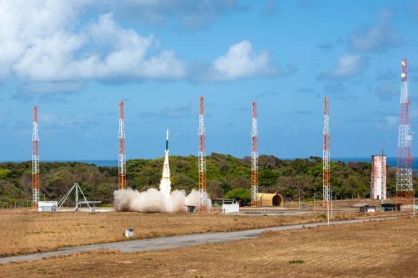 Foguete suborbital é lançado a partir do Centro de Lançamento da Barreira do Inferno (CLBI), em Parnamirim (RN). A imagem mostra o foguete em ascensão, com uma nuvem de fumaça na base, cercado pelas torres e estruturas da plataforma de lançamento, sob céu azul com nuvens.
