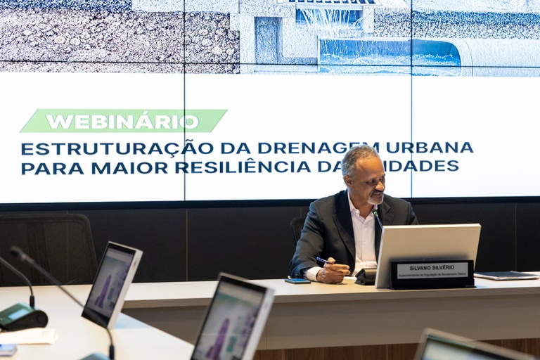 Gestão de riscos e planejamento das cidades
Em sua apresentação sobre a reconstrução das cidades a partir dos sistemas de drenagem, o professor Nilo Nascimento, da Universidade Federal de Minas Gerais (UFMG), destacou três frentes fundamentais para a gestão de riscos: conhecimento do risco, desenvolvimento de planos de prevenção e elaboração de planos de contingência. Segundo o professor, conhecer os riscos exige não apenas estudos hidrológicos, mas também uma avaliação ampla das consequências das inundações.
Nilo também chamou atenção para a necessidade de considerar a percepção das próprias comunidades sobre os riscos e o conhecimento desenvolvido por quem convive com esses eventos. “Há uma questão importante nesses estudos também, que é como a população percebe esse risco e como ela se organiza. É bem sabido que, mesmo quando há carências nas políticas públicas, a sociedade se organiza, as pessoas afetadas se organizam”, destacou. Segundo ele, o conhecimento acumulado pelas comunidades “tanto sobre o evento quanto sobre a forma de fazer face a ele é importante”.
Mudanças climáticas, NR nº 12 e soluções baseadas na natureza
A programação técnica também contou com apresentação do especialista em Recursos Hídricos e Saneamento Básico da ANA Lucas Cordeiro, da Superintendência de Estudos Hídricos e Socioeconômicos (SHE), sobre mudança do clima, recursos hídricos e diretrizes para adaptação.
A experiência da regulação após eventos extremos e a aplicação da NR nº 12/2025 foram abordadas por Tiago Luis Gomes, diretor-presidente da Agência Reguladora Intermunicipal de Saneamento do Rio Grande do Sul (AGESAN-RS), e Vagner Gerhart Mancio, diretor de Normatização da instituição. Priscilla Moura, da UFMG, apresentou como a norma trata a infraestrutura de drenagem.
O encontro também abordou a Estratégia Nacional de Soluções Baseadas na Natureza (ENSBN), em apresentação de Salomar Mafaldo de Amorin Junior, chefe do Departamento de Meio Ambiente Urbano do Ministério do Meio Ambiente e Mudança do Clima (MMA). Ele anunciou que a ENSBN será lançada em novembro, durante a 31ª Conferência das Nações Unidas sobre Mudanças Climáticas (COP31).
Na sequência, Eliana Teles do Carmo, coordenadora substituta de Capacitação do Sistema Nacional de Gerenciamento de Recursos Hídricos (SINGREH), apresentou o curso de Educação a Distância (EaD) sobre a NR nº 12/2025.
Moderado por Ana Cristina Strava, coordenadora de Regulação de Drenagem e Manejo de Águas Pluviais Urbanas da ANA, o webinário foi encerrado com espaço destinado ao debate e às perguntas do público.