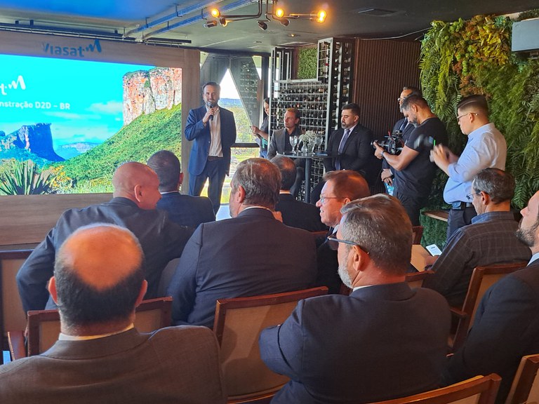 Presidente da Anatel, Carlo Baigorri, fala em frente a uma tela da Viasat, com plateia e equipe de filmagem presentes.