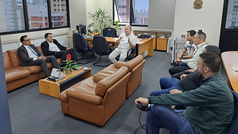Um grupo de homens em uma sala de escritório, em uma reunião. Eles estão sentados em dois sofás de couro marrom, enquanto um homem de camisa branca, no centro, gesticula enquanto fala. Há mesas de escritório ao fundo com computadores e outros equipamentos. Através das janelas, é possível ver outros prédios da cidade.
