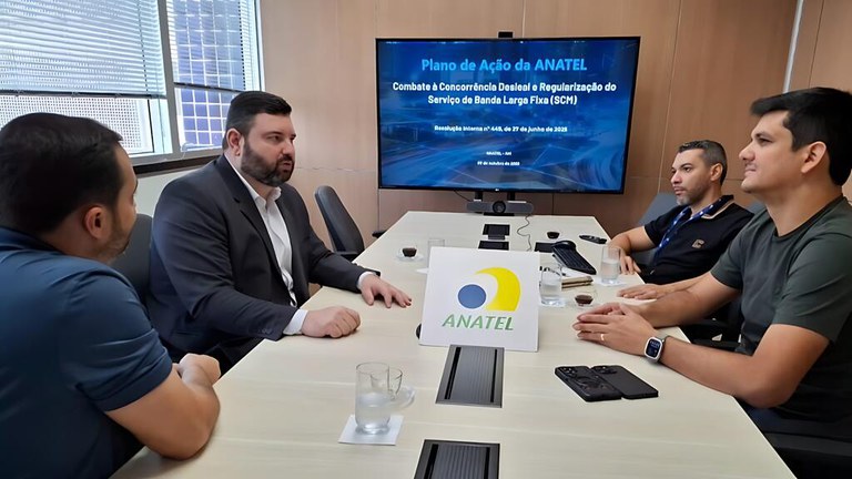 Quatro homens sentados em uma mesa de conferência com o logotipo da Anatel na mesa e um monitor mostrando uma apresentação da Anatel ao fundo.