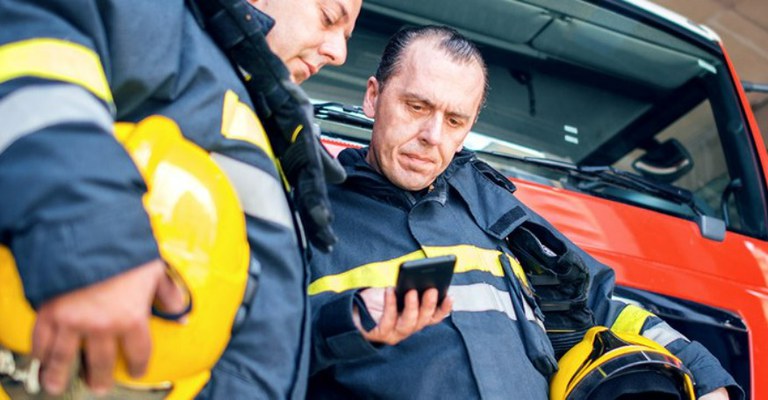 Na imagem há dois bombeiros, um deles olha para o celular que o outro está segurando.