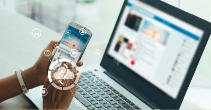 Em segundo plano uma mão segura um celular à frente de um laptop, em primeiro plano a imagem de uma "nuvem" remete a esse tipo de armazenamento de dados