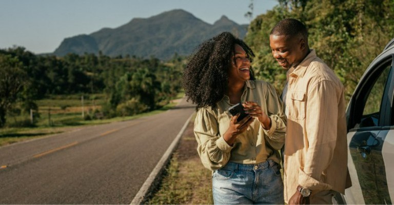 Na imagem, casal sorri ao lado do carro em uma estrada cercada por vegetação, e montanhas são visíveis no horizonte. Ambos sorriem enquanto a mulher segura um celular e o homem olha para o aparelho.