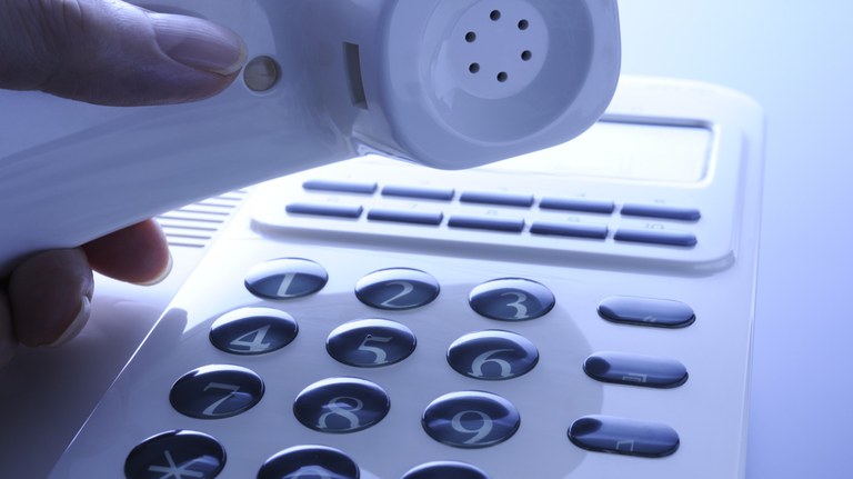 Uma mão segura o monofone de um telefone fixo branco, com o teclado numérico em foco.