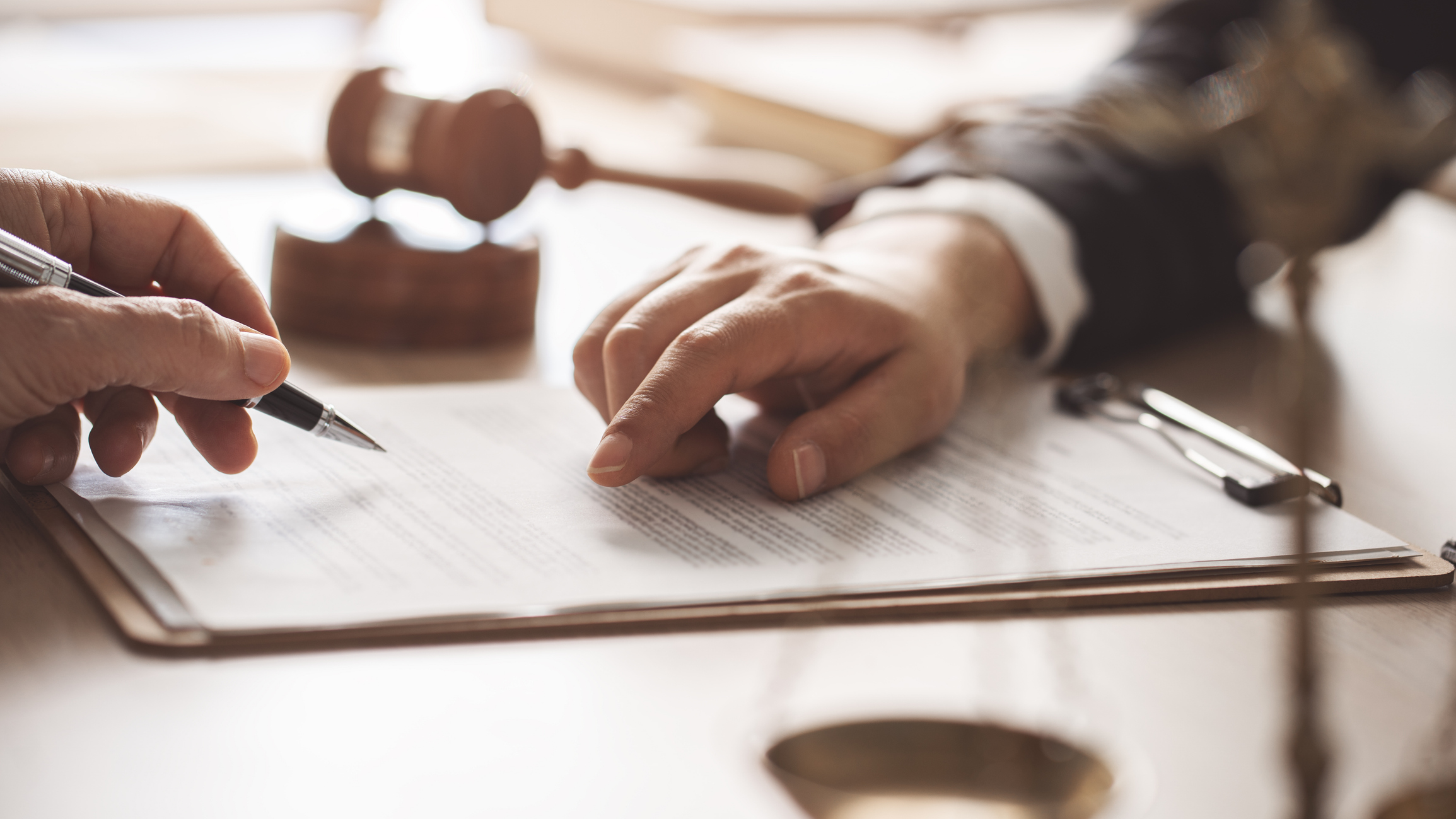 Duas pessoas estão revisando um documento legal em uma mesa. Uma mão aponta para uma seção do texto, enquanto a outra segura uma caneta para assinar. Ao fundo, fora de foco, há um martelo de juiz, simbolizando o contexto jurídico.