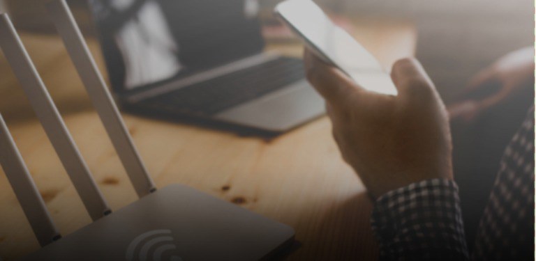 Imagem destaca uma mão com celular e um laptop e um roteador para banda larga sem fio em uma mesa