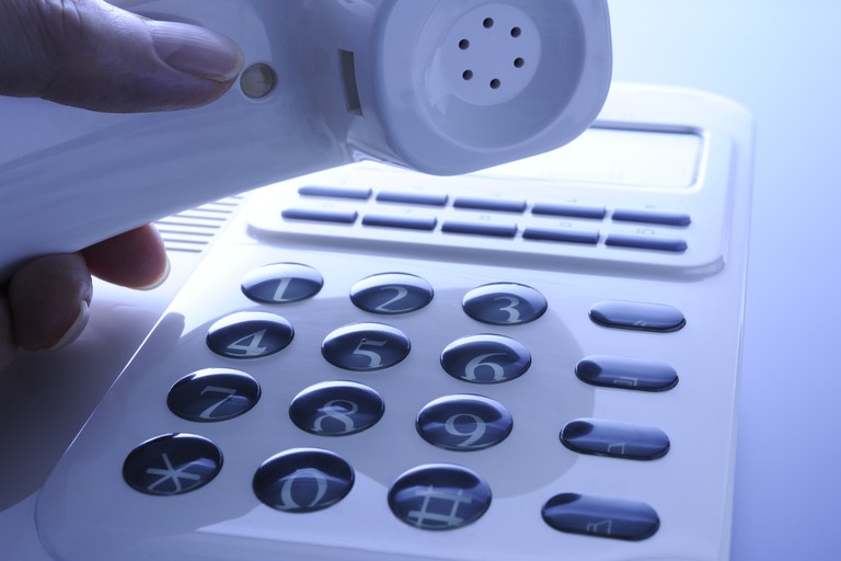 Uma mão segura o fone branco de um telefone fixo sobre o teclado numérico, indicando o ato de fazer uma ligação.