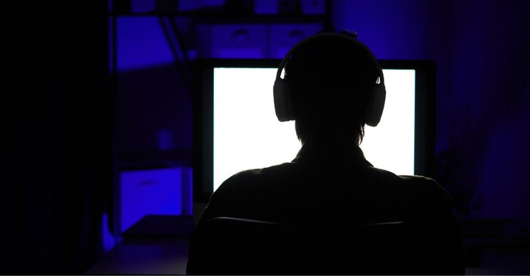 A imagem mostra a silhueta de uma pessoa sentada em frente a um monitor de computador em um ambiente escuro.