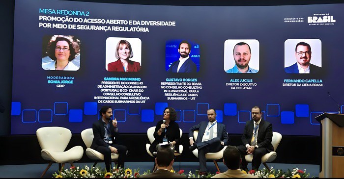 A imagem mostra quatro palestrantes sentados em um palco, com o superintendente-executivo da Anatel, Gustavo Borges, na extrema esquerda, falando ao microfone. Ele participa do Workshop Internacional de Cabos Submarinos e Data Centers, realizado em Brasília e promovido pelo Ministério das Comunicações. Um grande telão ao fundo exibe o título "MESA REDONDA 2: PROMOÇÃO DO ACESSO ABERTO E DA DIVERSIDADE POR MEIO DE SEGURANÇA REGULATÓRIA" e fotos e nomes dos cinco palestrantes, incluindo o dele. Também no telão, no canto superior direito, estão os logos do "MINISTÉRIO DAS COMUNICAÇÕES" e "BRASIL: UNIÃO E RECONSTRUÇÃO". Arranjos florais coloridos decoram a frente do palco. A plateia é vista em primeiro plano, de costas.