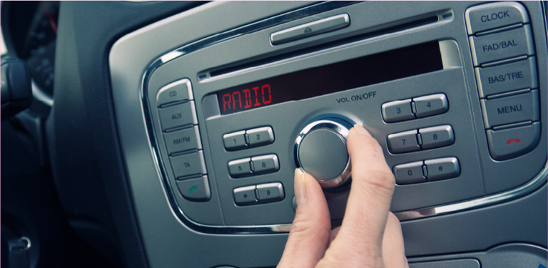 Em um carro, uma mão segura o volante e a outra mexe no botão do rádio. No dial do aparelho está escrito Rádio.