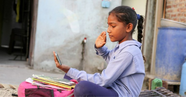 A imagem mostra uma menina sentada do lado de fora de uma casa, em um ambiente simples. Na sua frente há uma mochila rosa com alguns cadernos e livros em cima. A menina está segurando um celular com uma das mãos.