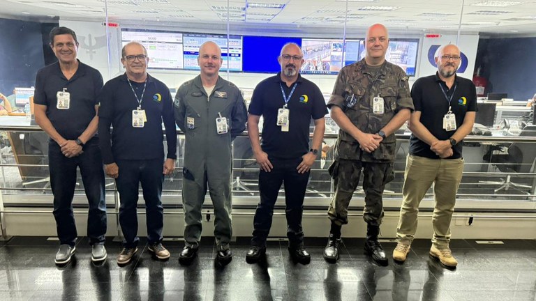 Esta imagem é uma fotografia em plano médio de seis homens adultos de pé lado a lado, posando para a câmera em uma sala de controle. Eles estão em um piso preto brilhante e reflexivo. Atrás deles, há uma divisória de vidro que separa a área onde estão de um centro de operações mais amplo, com mesas, cadeiras e várias telas grandes exibindo gráficos, dados e imagens de câmeras. A iluminação é artificial, proveniente de lâmpadas embutidas no teto.