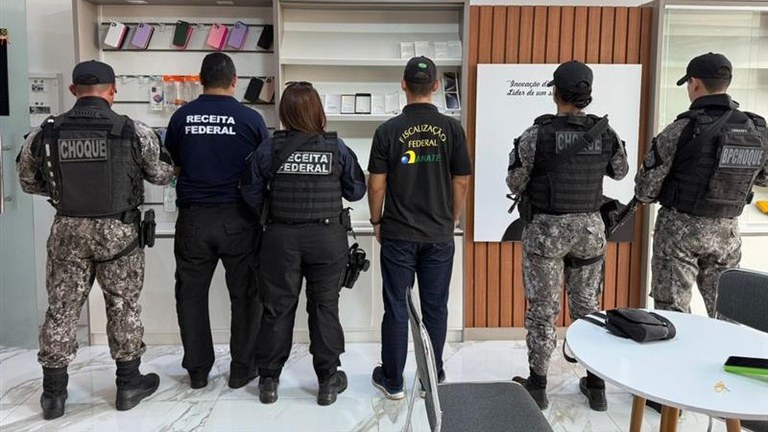 Agentes de Segurança e Fiscalização: Seis agentes estão de costas, observando prateleiras com produtos (como capas de celular e caixas de aparelhos).