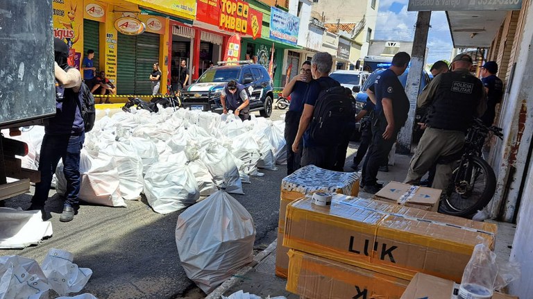 Uma fotografia de uma operação fiscal da Receita Federal no Brasil, com dezenas de grandes sacos brancos cheios de mercadorias no meio da rua, ao lado de um caminhão. As ruas de uma cidade movimentada podem ser vistas ao fundo. Caixas de papelão estão espalhadas na calçada, e agentes de segurança e policiais militares estão em pé na calçada. Uma viatura da polícia, um carro da Receita Federal e uma motocicleta também podem ser vistos. A área está bloqueada por fita amarela e preta.