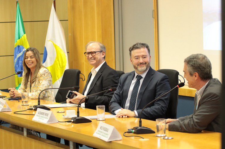 Fotografia colorida horizontal mostrando quatro pessoas sentadas lado a lado atrás de uma longa mesa de madeira clara, em um ambiente de conferência. Todas estão sorrindo e parecem interagir entre si. Ao fundo, no canto esquerdo, estão hasteadas a bandeira do Brasil e uma bandeira branca com um logotipo em azul e amarelo.  Identificação das pessoas da esquerda para a direita:  Ana Veneroso, representante da UIT para as Américas, uma mulher de pele clara, cabelos longos e lisos, vestindo um blazer estampado em tons claros.  Márcio Muniz de Farias, vice-reitor da UnB, um homem de pele clara, cabelos grisalhos curtos e óculos, vestindo terno preto e camisa branca.  Alexandre Freire, conselheiro, um homem de pele clara, barba cerrada e cabelos escuros, vestindo terno azul-marinho e gravata de pois.  Nilo Pasquali, superintendente de Planejamento e Regulamentação, um homem de pele clara e cabelos grisalhos, visto de perfil, vestindo terno cinza.