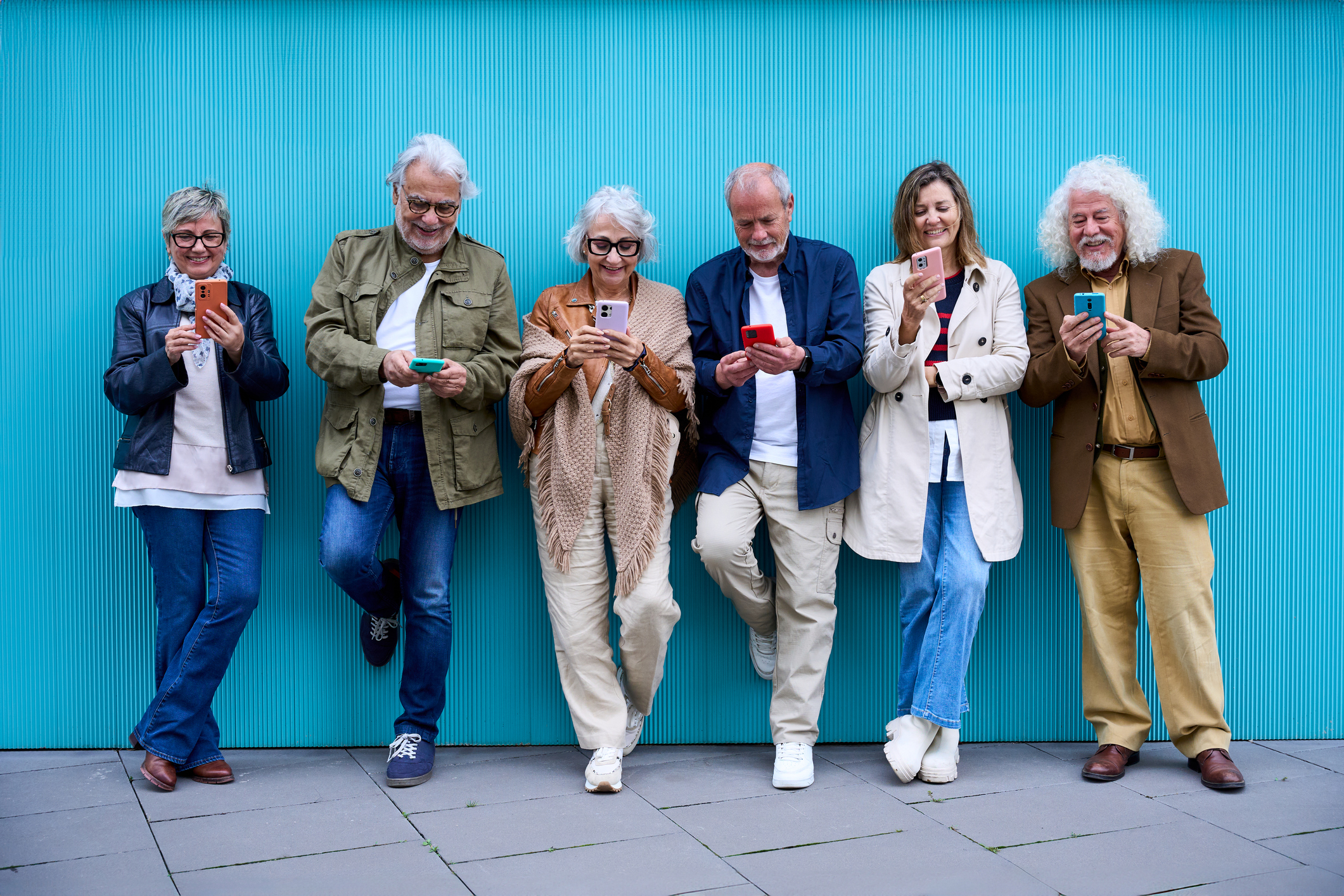 Grupo de idosos usando dispositivos tecnológicos juntos online, encostados em uma parede azul. Amigos maduros e alegres se divertindo assistindo redes sociais em celulares. Tecnologia e conceito moderno e antigo