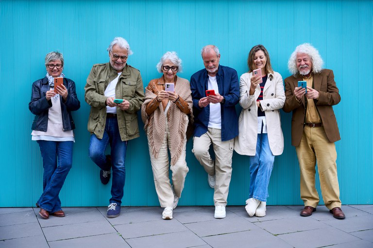 Grupo de idosos usando dispositivos tecnológicos juntos online, encostados em uma parede azul. Amigos maduros e alegres se divertindo assistindo redes sociais em celulares. Tecnologia e conceito moderno e antigo