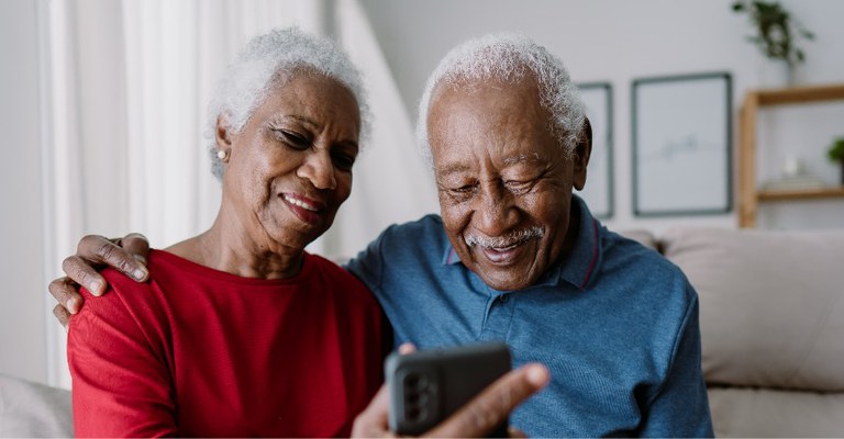 A imagem mostra um casal de idosos sorrindo enquanto olha para a tela de um smartphone.
