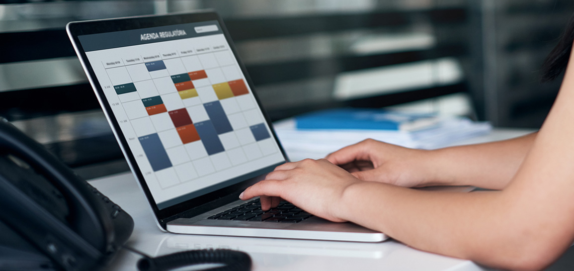 Na imagem são visíveis os braços e as mãos de uma mulher que digita em laptop com um calendário na tela, na mesa, à esquerda, há um telefone.