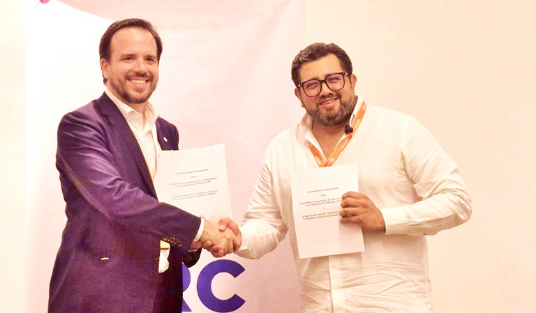 Na foto aparecem dois homens em um ambiente de evento institucional, apertando as mãos e sorrindo para a câmera. Ambos seguram documentos impressos, sugerindo a assinatura de um memorando.