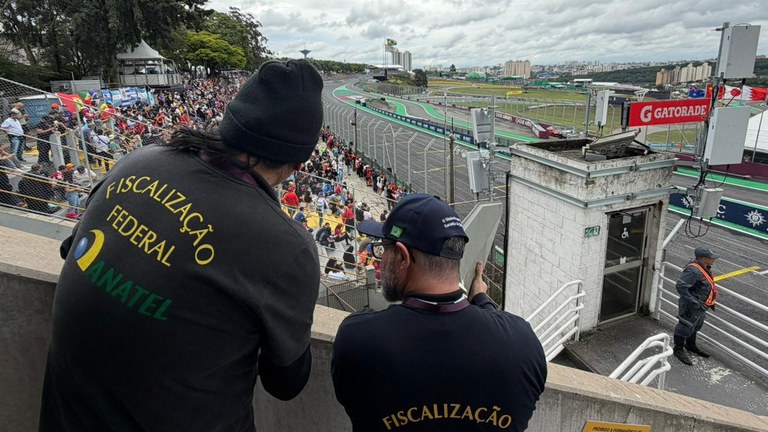 Dois homens estão de costas para a câmera, olhando para uma pista de corrida movimentada e arquibancadas cheias. O homem da esquerda usa um moletom preto com as palavras "FISCALIZAÇÃO FEDERAL" e "ANATEL" em amarelo e verde. O homem da direita usa um boné de beisebol e uma camiseta preta com a palavra "FISCALIZAÇÃO" nas costas. O dia está nublado, e a pista de corrida tem bandeiras e outdoors, incluindo um com o logotipo "GATORADE". À direita, um guarda de segurança veste um colete reflexivo.