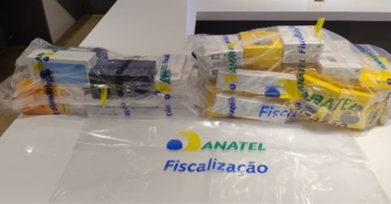 Produtos eletrônicos irregulares, acondicionados e lacrados em sacos transparentes sobre uma mesa, onde também é visível o logotipo institucional da entidade fiscalizadora, Anatel.