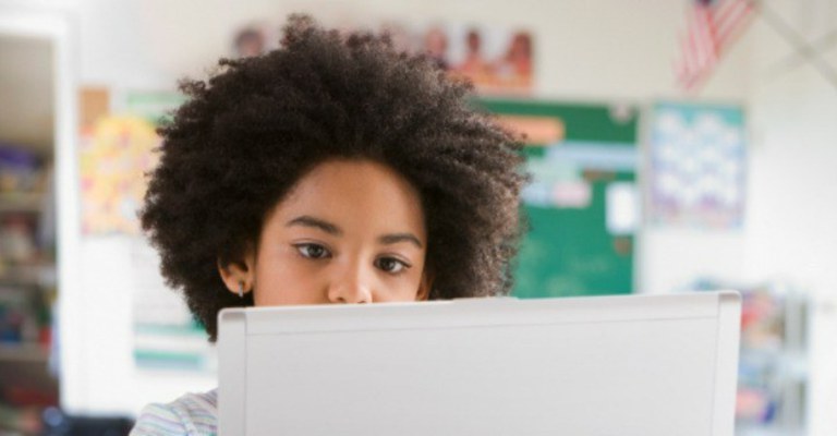 A imagem mostra uma criança com cabelos cacheados e volumosos, focada em um dispositivo eletrônico, possivelmente um laptop ou tablet branco, que está à sua frente. O fundo sugere um ambiente escolar: há um quadro verde e cartazes coloridos na parede.