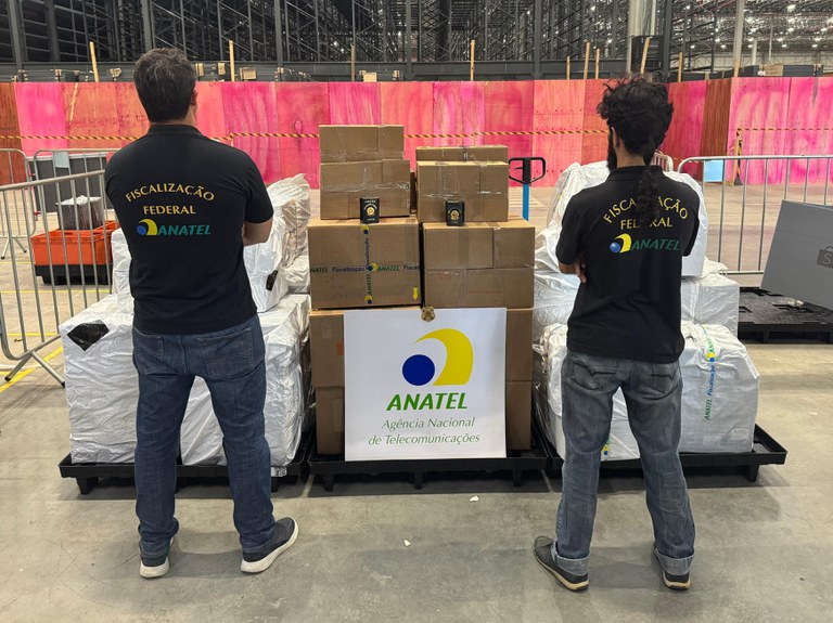 Dois homens com uniformes da ANATEL estão de costas, observando caixas de papelão e pacotes embalados em paletes pretos dentro de um armazém com fundo rosa e amarelo. Uma placa branca com o logotipo da ANATEL está na frente das caixas.