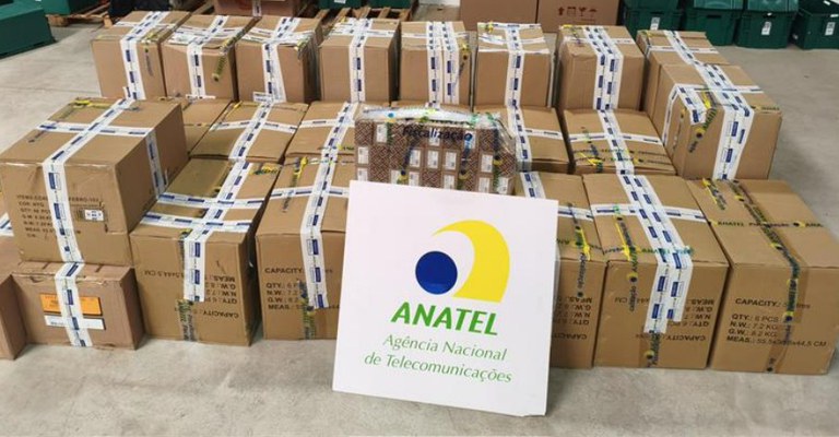 A imagem mostra um conjunto de caixas de papelão lacradas com fitas de segurança que possuem a marca da Anatel, indicando produtos irregulares de telecomunicações lacrados pela agência reguladora durante a Operação Produto Legal, 1º/12/2025.