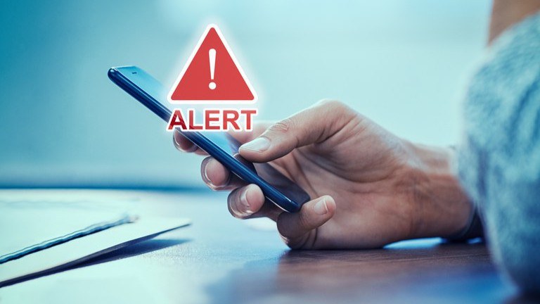 Uma imagem focada em uma mão segurando um smartphone. Na tela do celular, há uma sobreposição digital de um triângulo de aviso vermelho com um ponto de exclamação, e a palavra "ALERT" em letras vermelhas abaixo dele. O fundo está desfocado e mostra uma pessoa de costas, sentada em uma mesa, com alguns papéis.