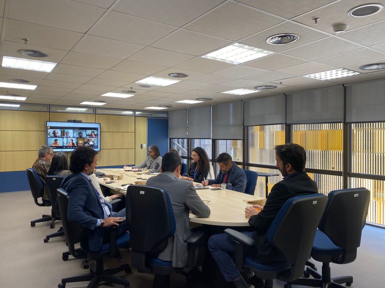 Reunião em uma sala de conferências, com algumas pessoas presentes na sala e outras participando por videochamada em uma tela grande.