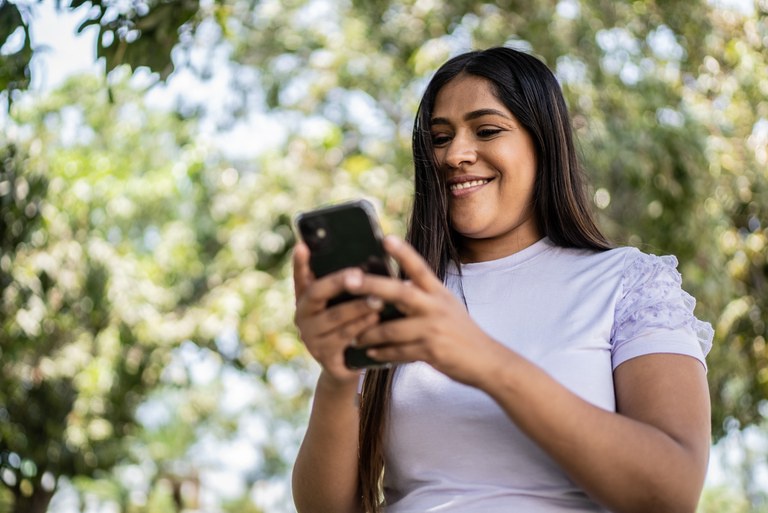 Mulher de pele morena segura um telefone celular em local aberto