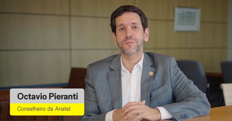 Octavio Pieranti, conselheiro da Anatel, aparece sentado à mesa em ambiente corporativo, falando diretamente à câmera em vídeo apresentado na abertura do Encontro da Rede Nacional de Comunicação Pública (RNCP) 2026; a imagem traz identificação em sobreposição com o nome e o cargo.