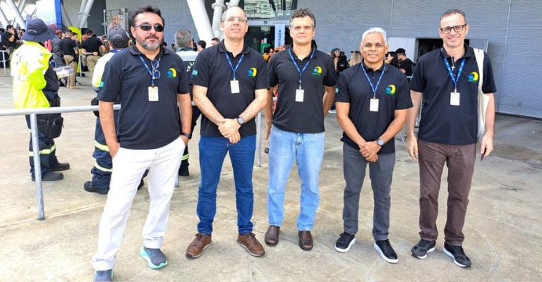 Cinco pessoas, todas identificadas como servidores da Anatel, posam lado a lado em área externa da Arena Castelão, em Fortaleza. Todas usam camisetas pretas com o logotipo da Agência e crachás de identificação. Ao fundo, há a estrutura moderna do estádio, com colunas e fachada metálica, além de profissionais de diferentes órgãos atuando em um cenário de simulação de emergência, alguns com coletes refletivos e equipamentos. O ambiente é aberto, durante o dia, e transmite a ideia de uma ação integrada e organizada de preparação para situações de crise.