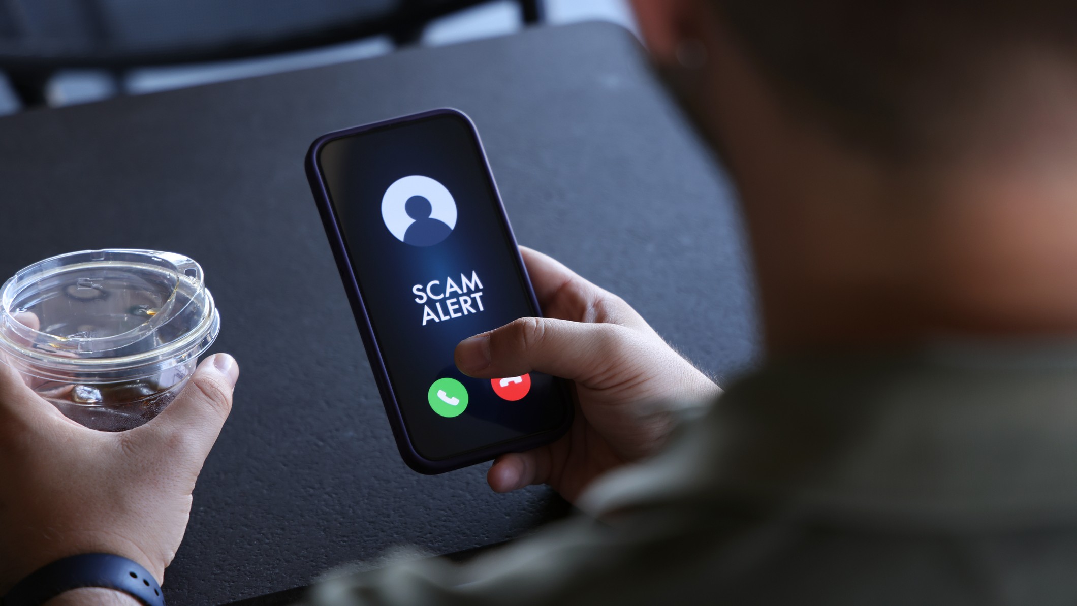 Um homem segura um smartphone que exibe um alerta de fraude ("SCAM ALERT") durante uma chamada recebida. Os botões para atender (verde) e recusar (vermelho) a chamada estão visíveis na parte inferior da tela. Na mão esquerda, ele segura um copo plástico transparente com gelo e um líquido claro, provavelmente água ou refrigerante. O fundo é desfocado, sugerindo um ambiente casual, como uma cafeteria, com uma mesa escura. A imagem ilustra o problema de chamadas fraudulentas e a importância de estar atento a alertas de segurança em dispositivos móveis.