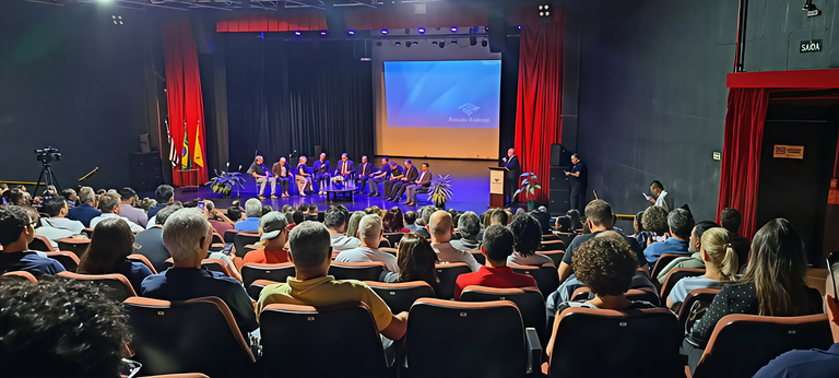 Um plano geral de um auditório lotado durante um evento. No palco, um grupo de oito homens sentados em cadeiras, com um homem em pé discursando num púlpito à direita e um grande telão projetando "Residência Educativa" atrás. Uma plateia numerosa ocupa as cadeiras do teatro. Cortinas vermelhas emolduram o palco. A iluminação é teatral, com luzes azuis sobre o palco.