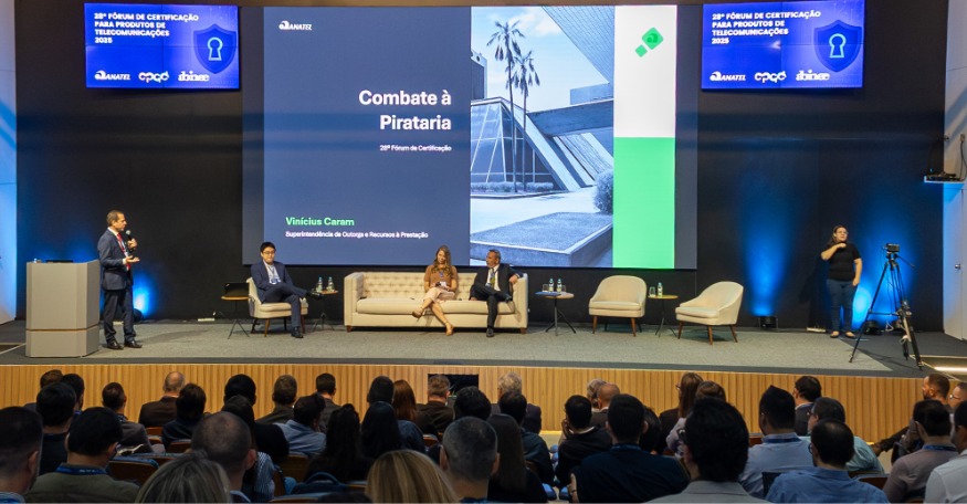 Auditório lotado com público atento a um painel no palco durante o 28º Fórum de Certificação em Campinas (SP). No centro, uma grande tela exibe o tema “Combate à Pirataria”. Três pessoas no palco estão sentadas em poltronas, enquanto outra apresenta em pé com microfone. No topo da tela, aparecem os logotipos da Anatel, CPqD e Abinee, reforçando a parceria institucional. O ambiente é moderno, com iluminação clara e clima profissional.