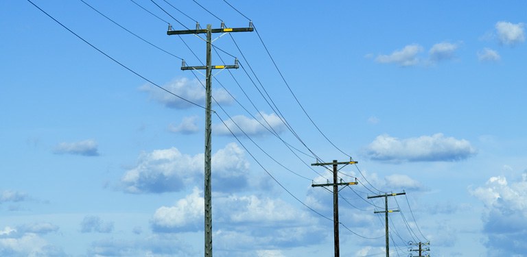 A imagem mostra uma fileira de postes de energia elétrica alinhados ao longo de uma via aberta, vistos em perspectiva.