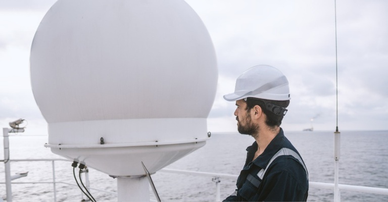 Técnico de serviços marítimos próximo a um terminal VSAT no convés de uma embarcação. Ele está trabalhando em um laptop.