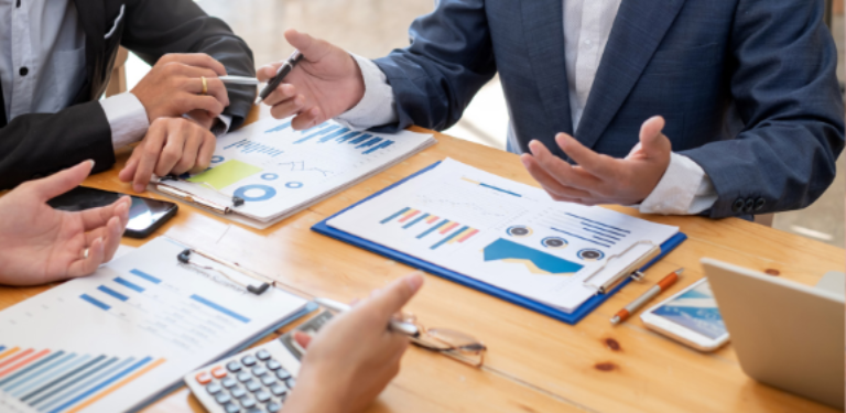 Pessoas em uma mesa de reunião com calculadora, celular, laptop e papéis com gráficos