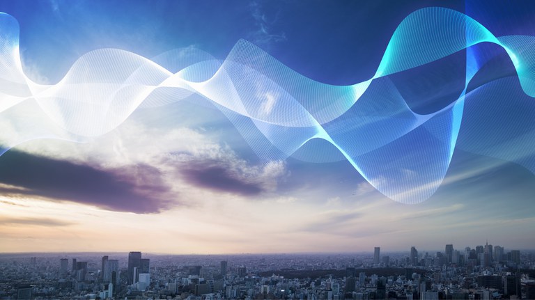 Um panorama urbano ao entardecer sob um céu que transita do azul ao roxo e rosa, com nuvens esparsas. Ondas de luz translúcidas, em tons de azul e branco, flutuam proeminentemente no céu, curvando-se elegantemente sobre os prédios da cidade.