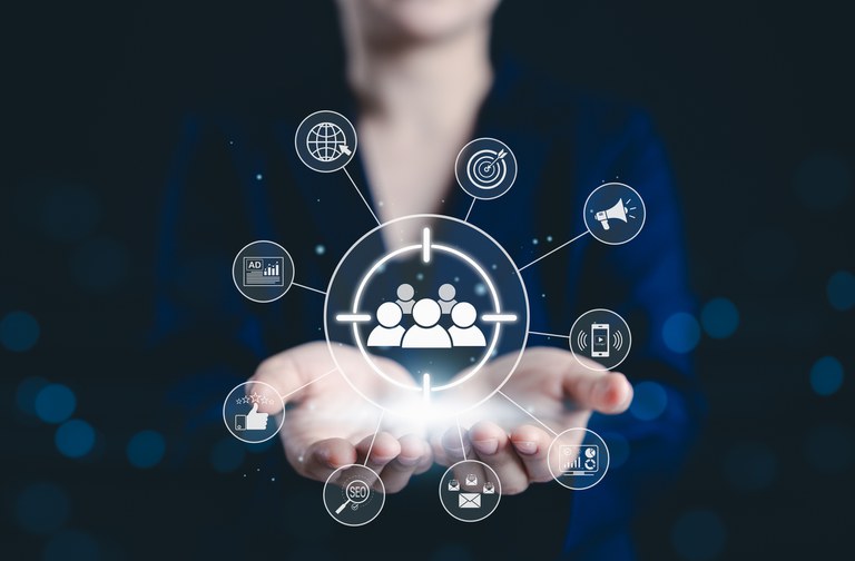 Mãos em postura de acolhimento sustentam um brilho central do qual emanam diversos ícones digitais, representando estratégias de marketing digital e o foco no público-alvo.