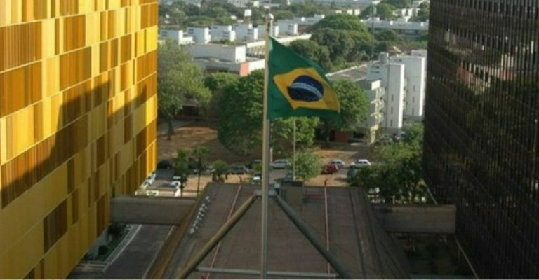 Bandeira do Brasil hasteada entre dois prédios da sede da Anatel em Brasília, um com fachada dourada e outro com fachada envidraçada escura. Ao fundo, área arborizada e construções residenciais.
