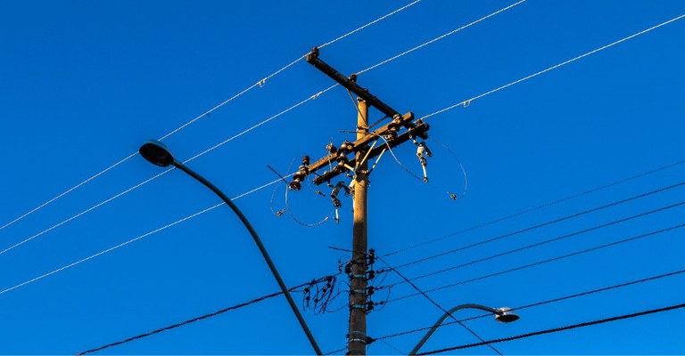 A foto mostra um poste, que pode ser usado para distribuição de serviços de energia elétrica e de telecomunicações, com fios e equipamentos de distribuição, contra um céu azul intenso e limpo.