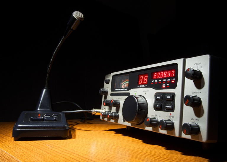 A imagem mostra um rádio transceptor de mesa e um microfone de pedestal, ambos posicionados sobre uma superfície de madeira clara. O ambiente ao redor está escuro, exceto pela área iluminada que destaca o equipamento.
