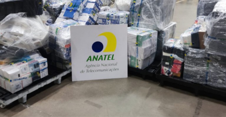 Placa com o logotipo da Anatel e o texto “Agência Nacional de Telecomunicações” posicionada à frente de pilhas de caixas embaladas em plástico.