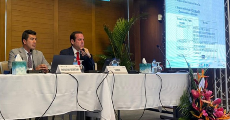 Vinicius Caram, à direita, preside a mesa de reunião da Citel ao lado de outro representante durante a 47ª Reunião do CCP.II, em Portsmouth, na Dominica. Ambos usam terno e gravata, sentados detrás de uma mesa com microfones, documentos e água. Ao fundo, há um telão com apresentação técnica.